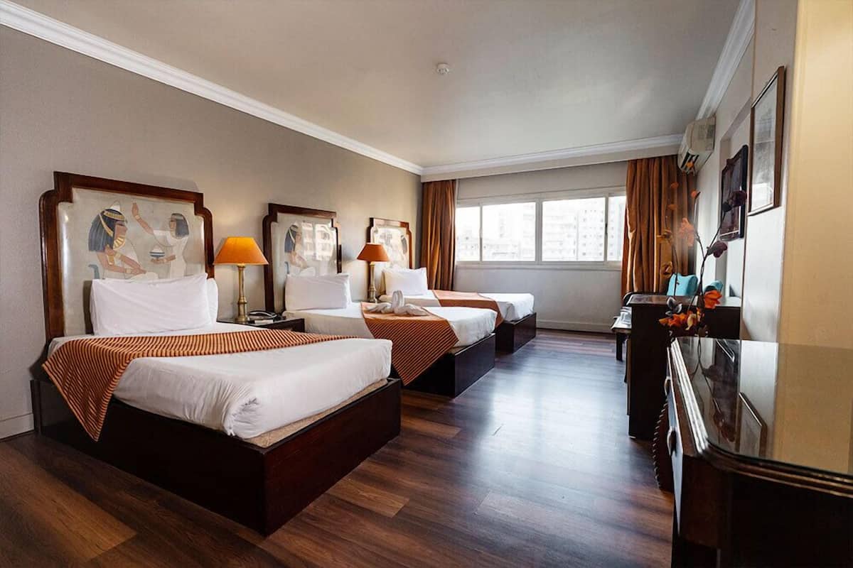 A hotel room with three beds, wooden flooring, a desk with a lamp, and artwork on the walls.