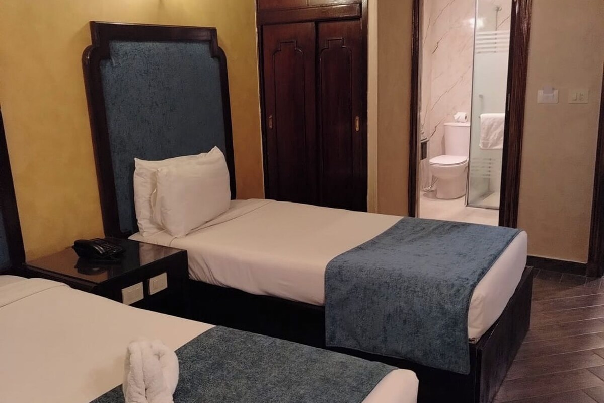 A hotel room with two single beds, a wooden wardrobe, a bathroom with a glass shower enclosure, and a bedside table with a phone.