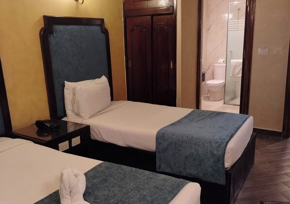 Habitación de hotel con dos camas individuales, un armario de madera, un baño con cabina de ducha de vidrio y una mesita de noche con teléfono.