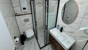 Un baño moderno con ducha de vidrio, piso de mármol, lavamanos blanco con espejo y toallero.