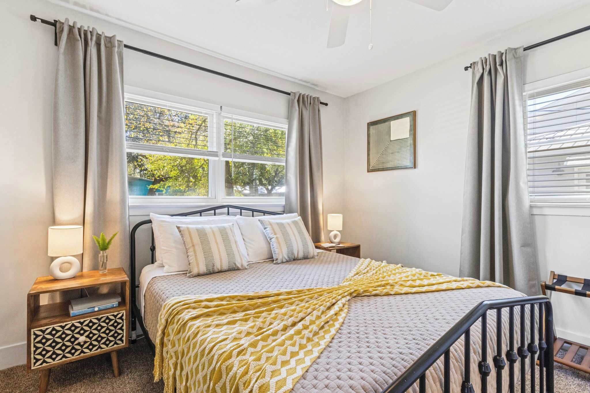 Une chambre à coucher comprenant un lit, des tables de chevet, une fenêtre avec des rideaux, un ventilateur de plafond et un miroir fixé au mur.