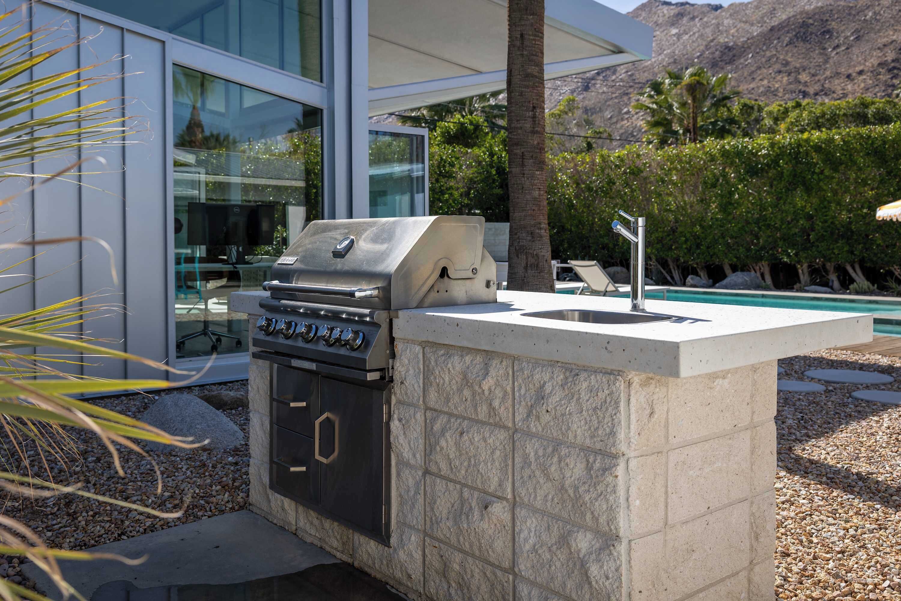 An outdoor kitchen with a grill, a sink, and a stone countertop, adjacent to a modern building with glass windows.