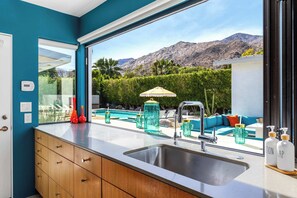 A modern kitchen with a large window, a built-in sink, and a view of a pool and mountains. - Luxury Home with Pool, Spa & Stunning Mountain Views (Palm Springs)