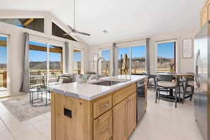 A modern kitchen with a large island, a dining area with a table and chairs, and large windows offering views of the outdoors.