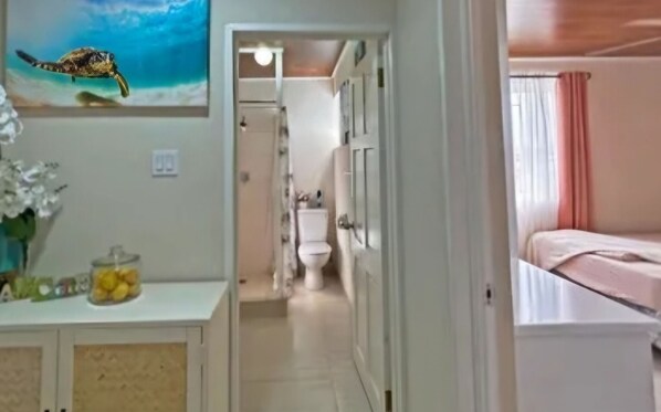 A hallway leading to a bathroom with a toilet and a sink. There is a bed with pink curtains on the right.