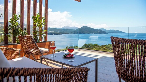 Une terrasse meublée de mobilier en osier, une table avec un verre de vin rouge et une vue imprenable sur la mer et les montagnes.