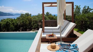 Un salon de bord de piscine doté d’une terrasse en bois, de chaises blanches rembourrées, d’une table avec des fruits et offrant une vue sur la mer et les montagnes.