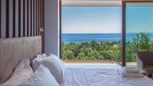 Une chambre avec un grand lit, du linge de lit blanc et une vue sur la mer et la verdure, visible à travers une porte vitrée.