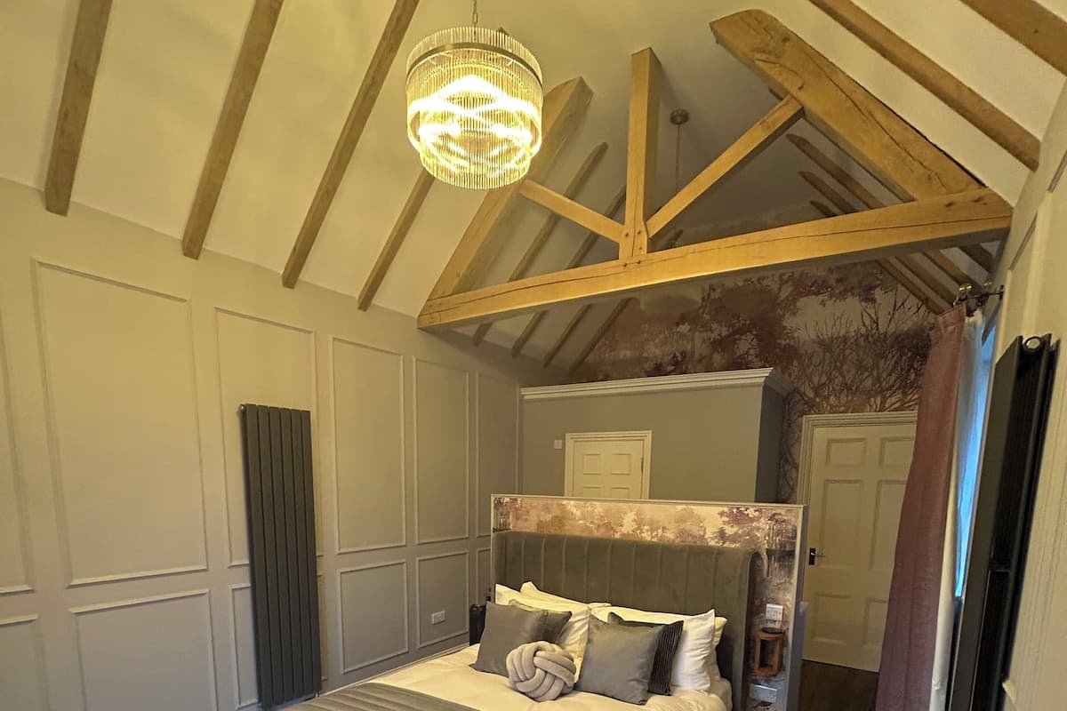 A bedroom with a large bed, wooden beams, a chandelier, and a view of the bathroom.