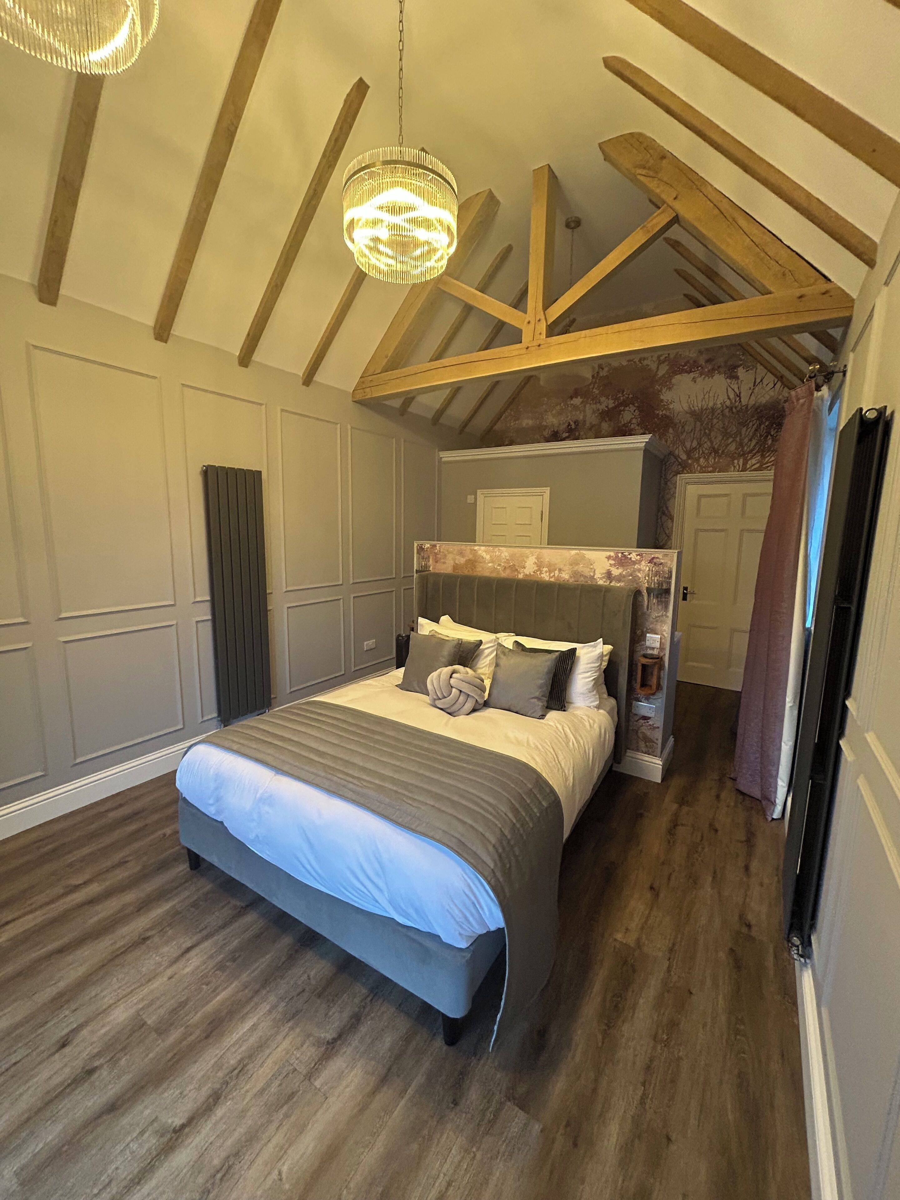 A bedroom with a large bed, wooden beams, a chandelier, and a view of a bathroom.