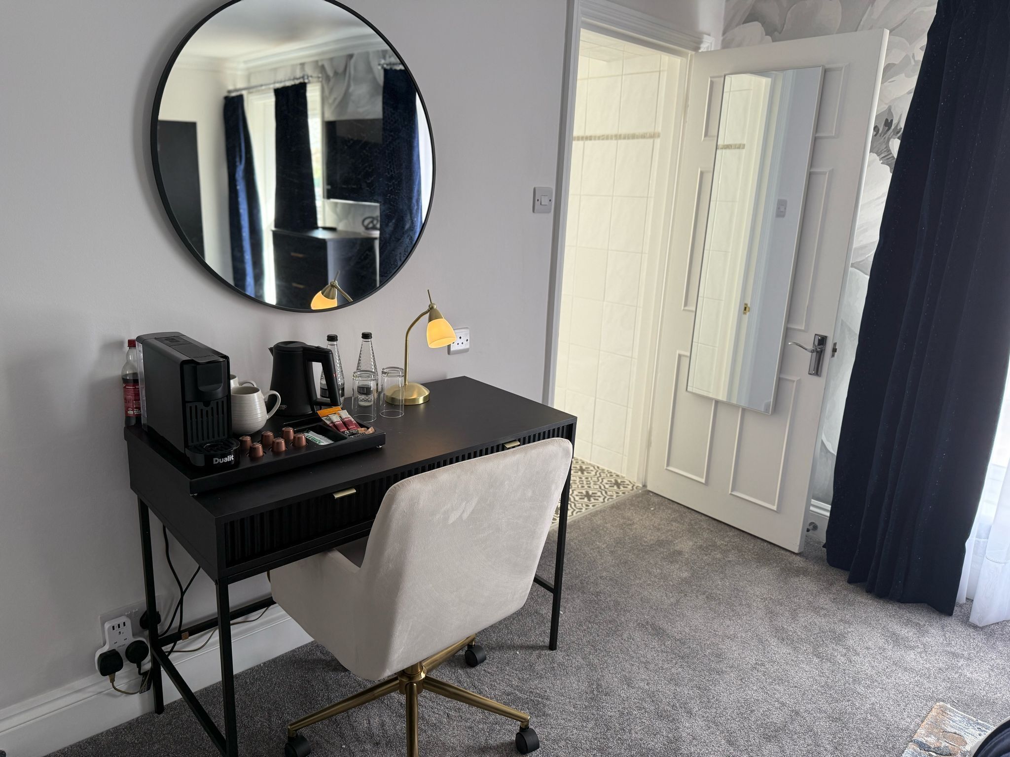 A hotel room with a black desk, a round mirror, a white chair, a coffee maker, and a lamp.