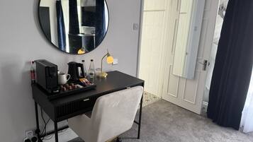 A hotel room with a black desk, a round mirror, a white chair, a coffee maker, and a lamp.
