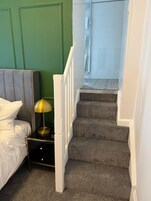 Un dormitorio con cama, mesita de noche, lámpara y una escalera que lleva a un baño.