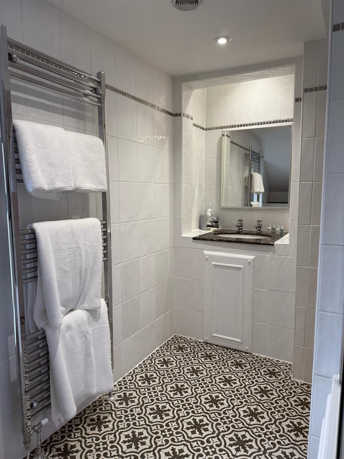 A modern bathroom with white tiles, a patterned floor, a glass shower enclosure, a sink with a mirror, and two towels on a towel rack.