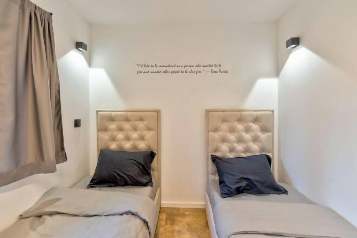 Una habitación pequeña y bien iluminada con dos camas, cada una con un cabecero tapizado y una almohada. Hay una cita en la pared.