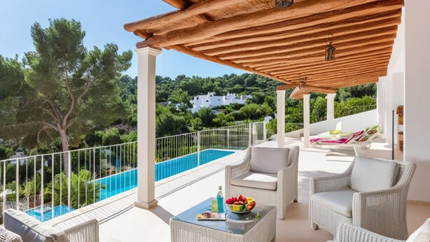 Un espacio al aire libre techado con mobiliario de mimbre, una piscina y vistas a zonas verdes y edificios al fondo.