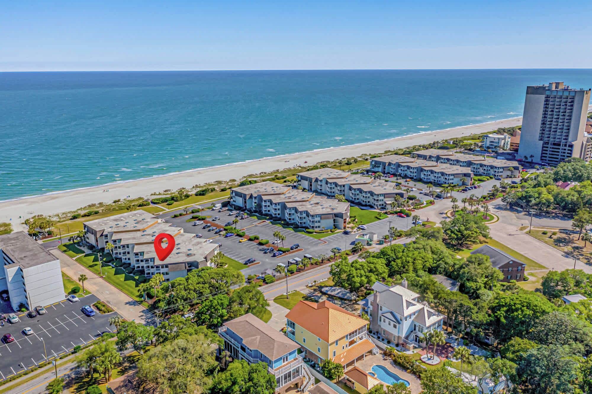 An aerial view of a coastal hotel with multiple buildings, a parking lot, and a beach in the background.