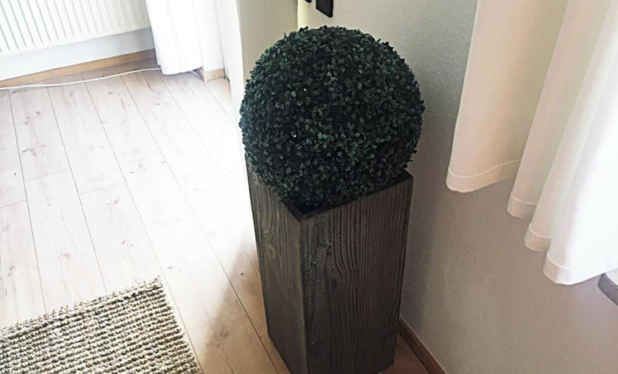 Um canto com piso de madeira, um cachepot de madeira escura com topo esférico e uma cortina branca.