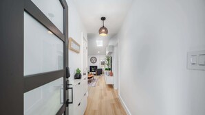 Un pasillo moderno con suelo de madera, una puerta de cristal, una lámpara colgante y un área de comedor con una mesa y sillas al fondo.