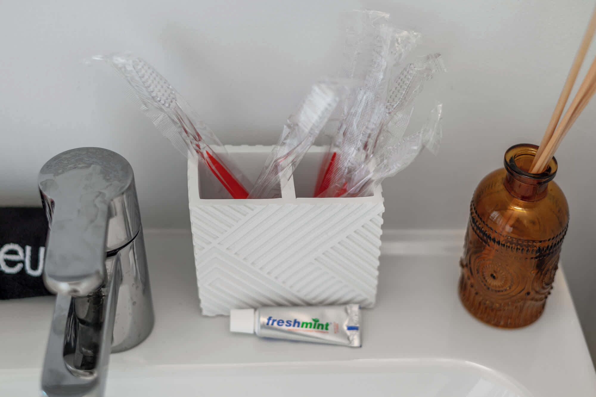 A bathroom sink with a toothbrush holder containing toothbrushes, a tube of freshmint toothpaste, and a decorative bottle.