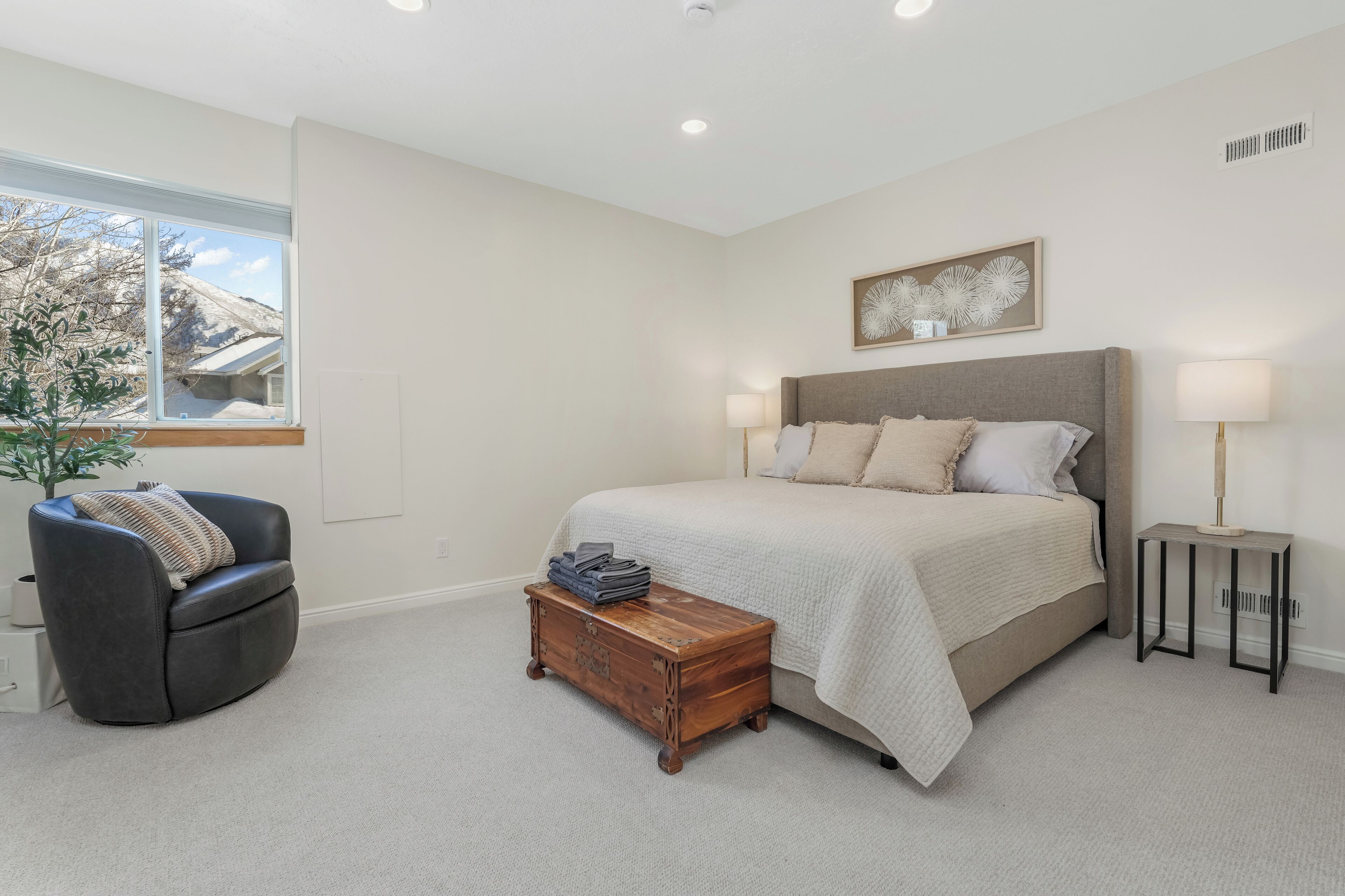 Een slaapkamer met een bed, nachtlampjes, een klein houten kistje, een zwarte fauteuil en een raam met uitzicht op een berg.