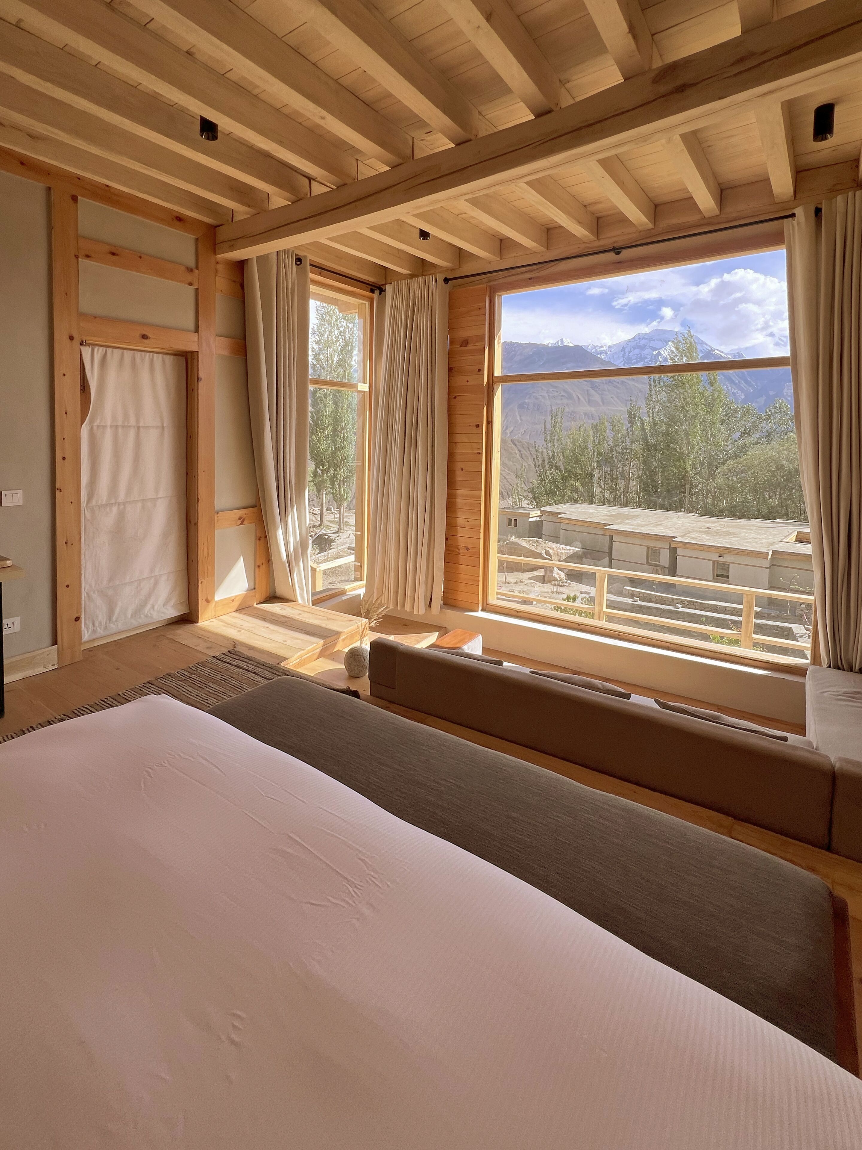 A bedroom with a large bed, wooden ceiling beams, a window with a view of mountains, and a window seat.