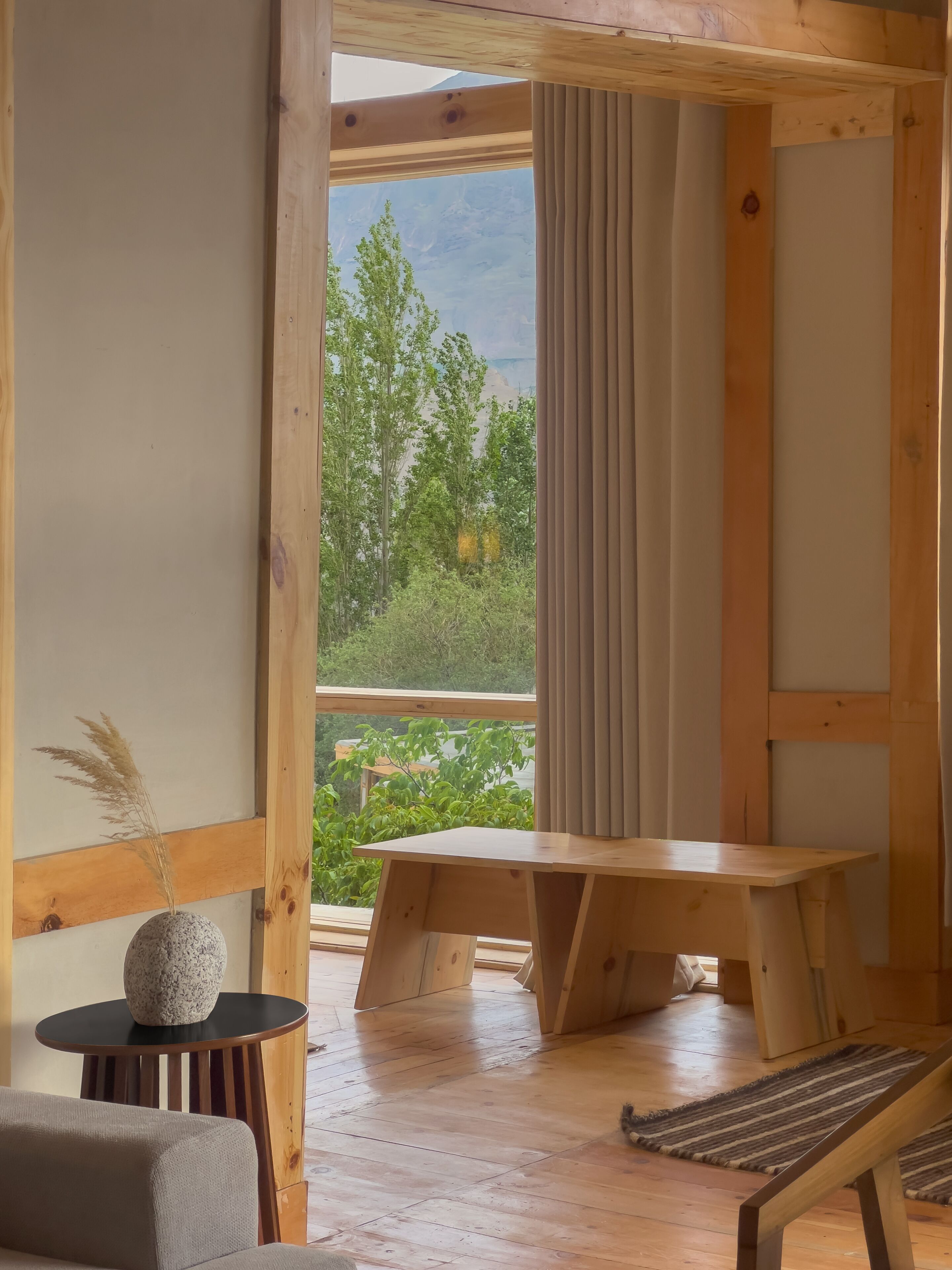A wooden interior with a large window, a stone vase with dried plants, a wooden bench, and a small table.