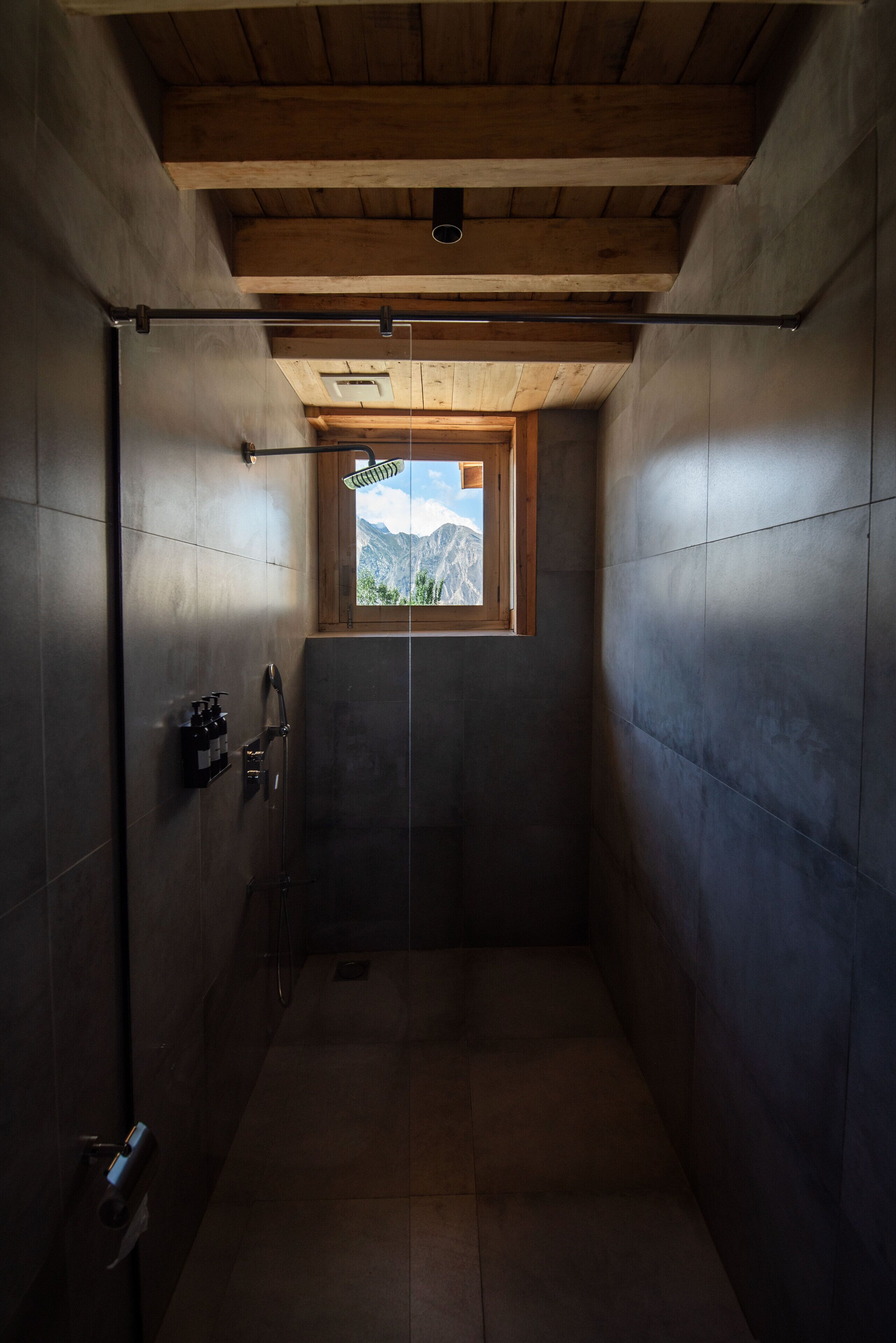 A narrow shower area with a window view of mountains, wooden beams on the ceiling, and a hand dryer on the wall.