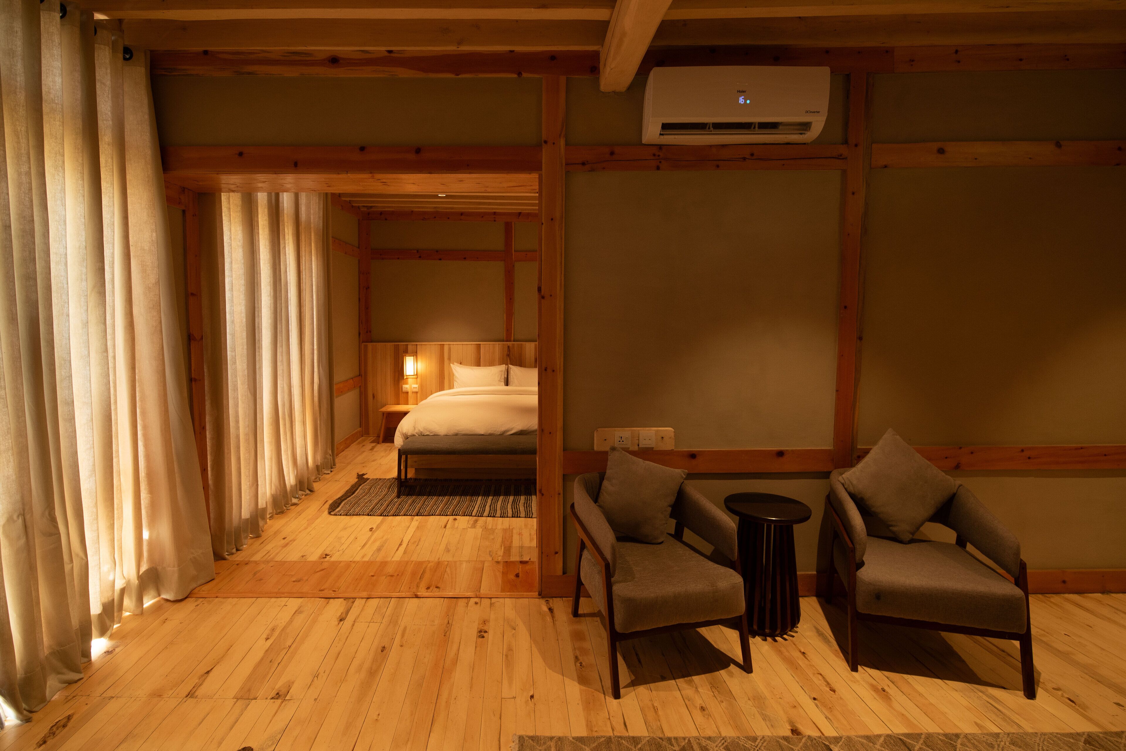 A wooden-floored room with a bed, two armchairs, a small table, and an air conditioning unit.