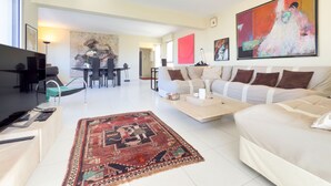 A modern living room with a sofa, a coffee table, and a TV. There is a dining area with a table and chairs. - L'Artiste (Cannes)