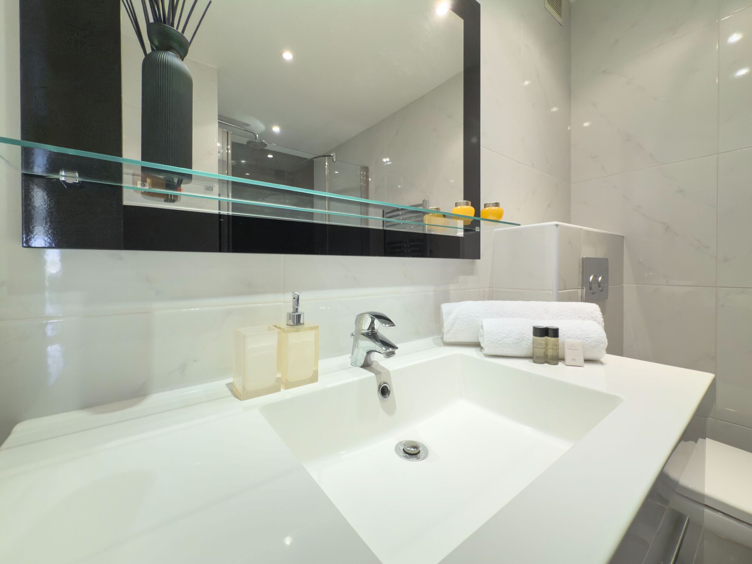 Un baño moderno con un lavamanos grande de color blanco, un estante de vidrio sobre el espejo y una toalla blanca en la pared.