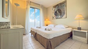 A hotel room with a large bed, a desk with a lamp, and a painting on the wall. - L'Artiste (Cannes)