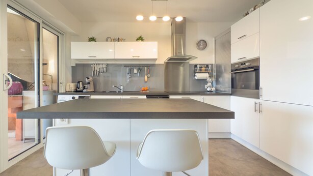 Een moderne keuken met witte kasten, een centraal eiland met twee barkrukken, roestvrijstalen apparatuur en een glazen spatwand.