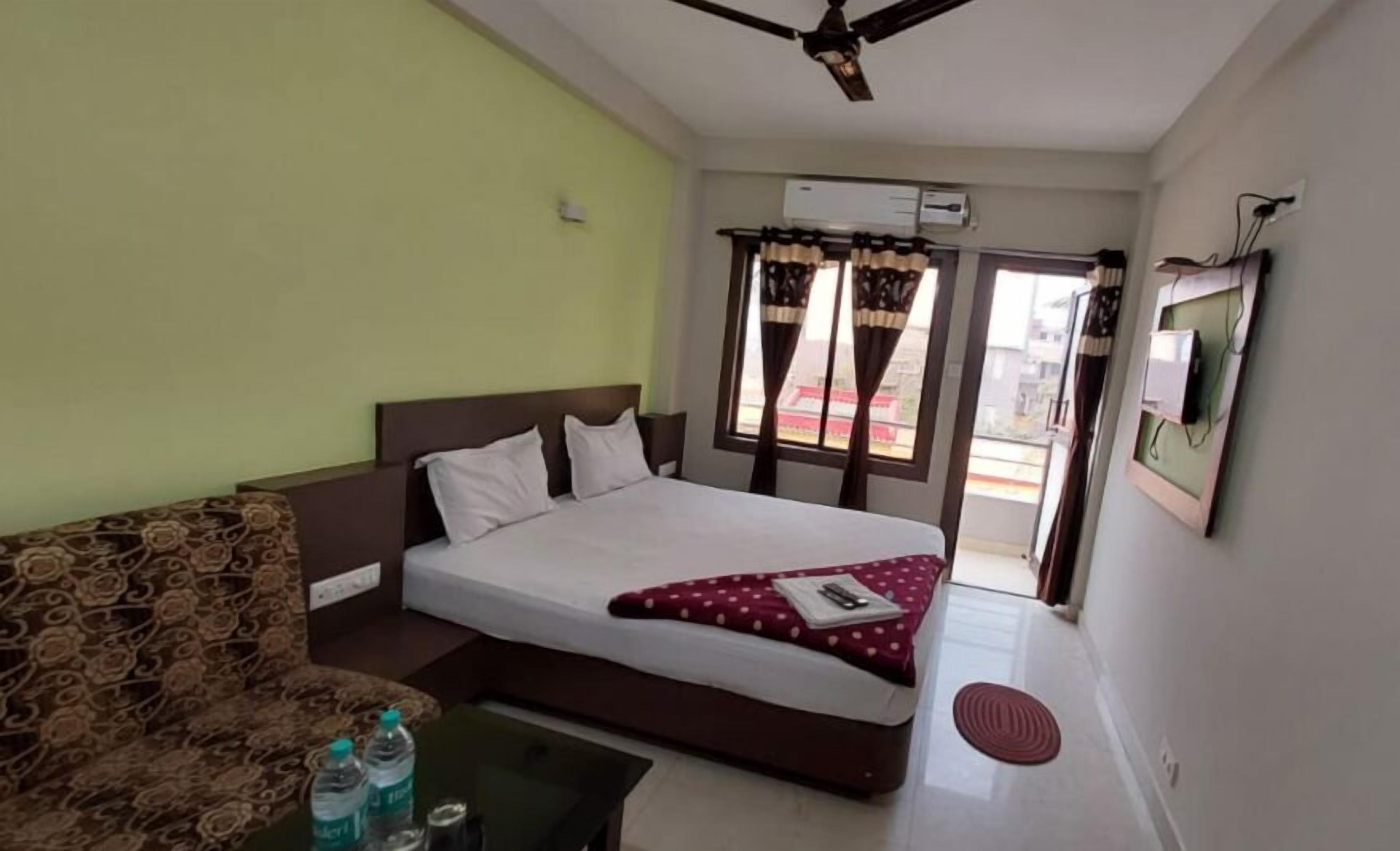A hotel room with a bed, a sofa, a coffee table with water bottles, a ceiling fan, and an air conditioning unit.