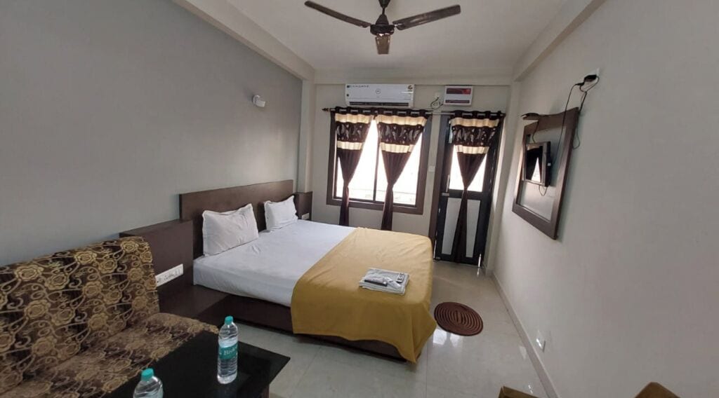 A hotel room with a bed, a sofa, a table with a water bottle, an air conditioning unit, and curtains.