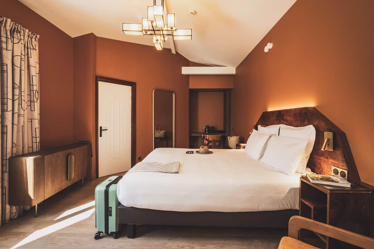 Habitación de hotel con una cama grande, una mesita de noche con lámpara, un armario, un espejo y una maleta.