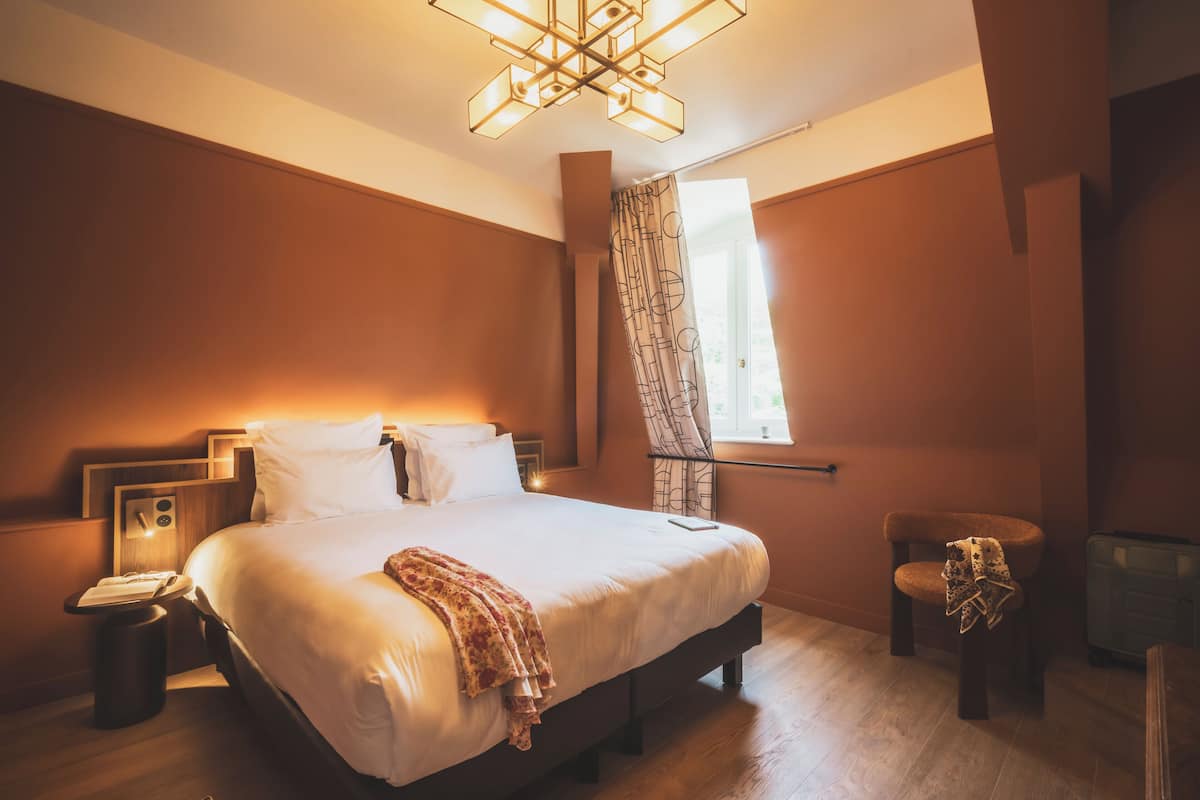 Un dormitorio con una cama grande, mesitas de noche, una lámpara de noche, una silla, una ventana con cortinas y una lámpara en el techo.
