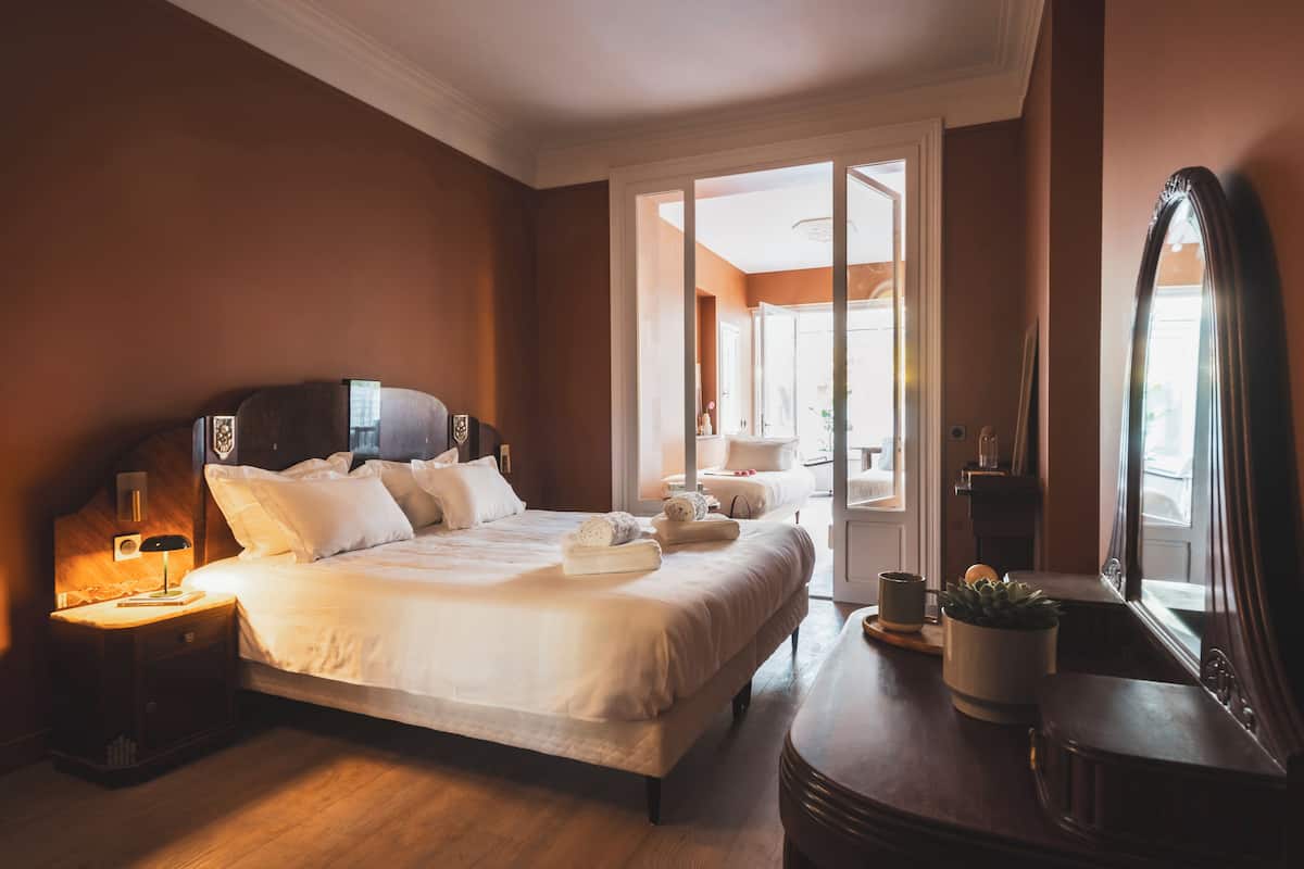 Habitación de hotel con una cama grande, mesita de noche, lámpara, espejo y vistas al exterior.