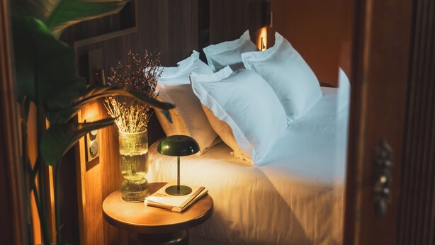 Una cama bien tendida con sábanas y almohadas blancas, una mesita de noche con una lámpara y un libro, y un jarrón con flores secas.