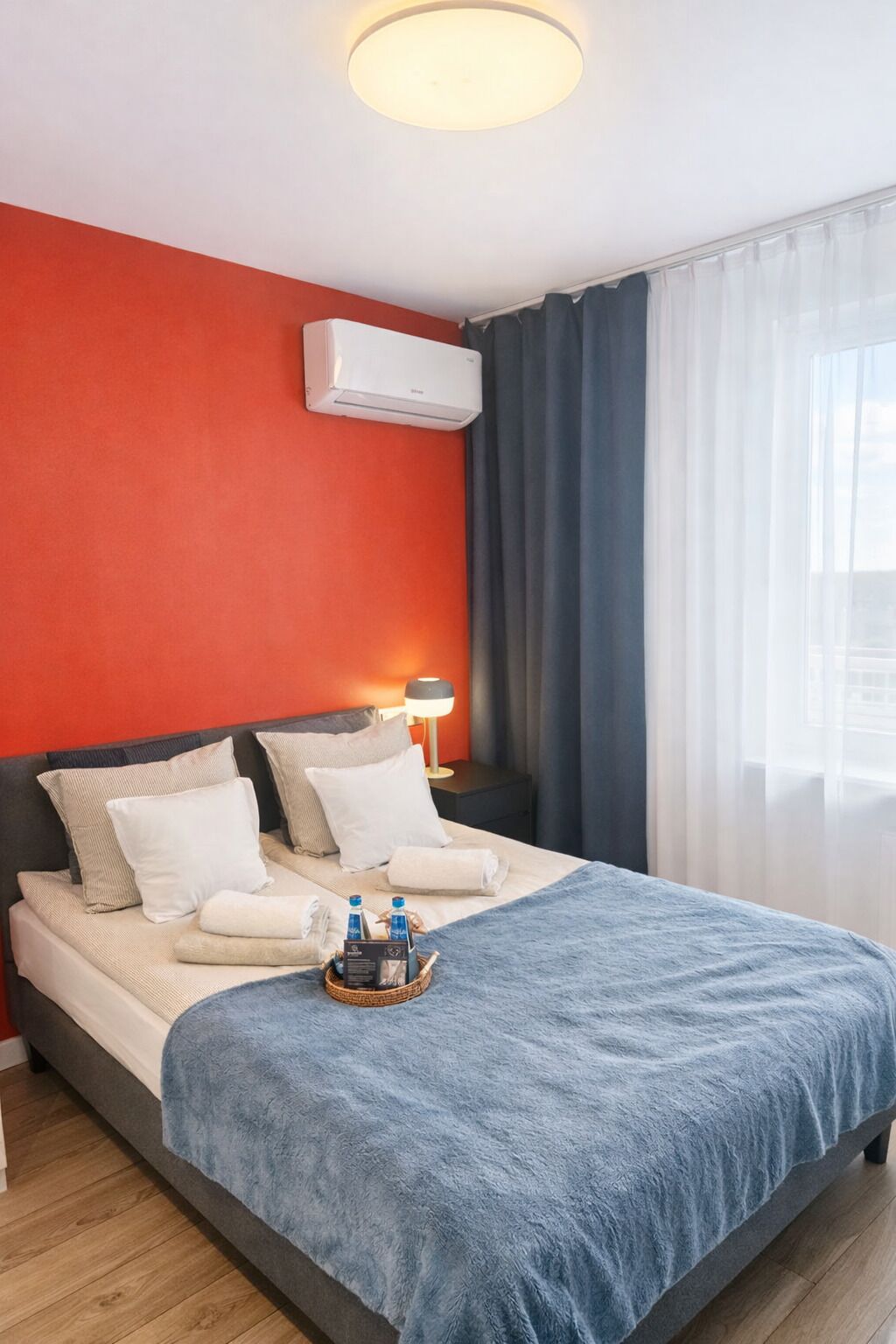 Habitación de hotel con cama, mesita de noche con lámpara, aire acondicionado y ventana con cortinas.