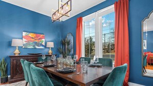 A dining area with a wooden table set for six, teal chairs, and a large window with red curtains. - Cosmopolitan- Hot Tub, Heated Pool & Fire Pit (Fredericksburg)