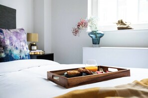 A neatly made bed with a breakfast tray containing muffins, a glass of orange juice, and cutlery. A vase with flowers and a lamp are visible in the background.