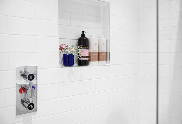 A bathroom with white tiled walls, a shower with a chrome faucet, and a shelf with toiletries including a black bottle, a pink bottle, and two white bottles.