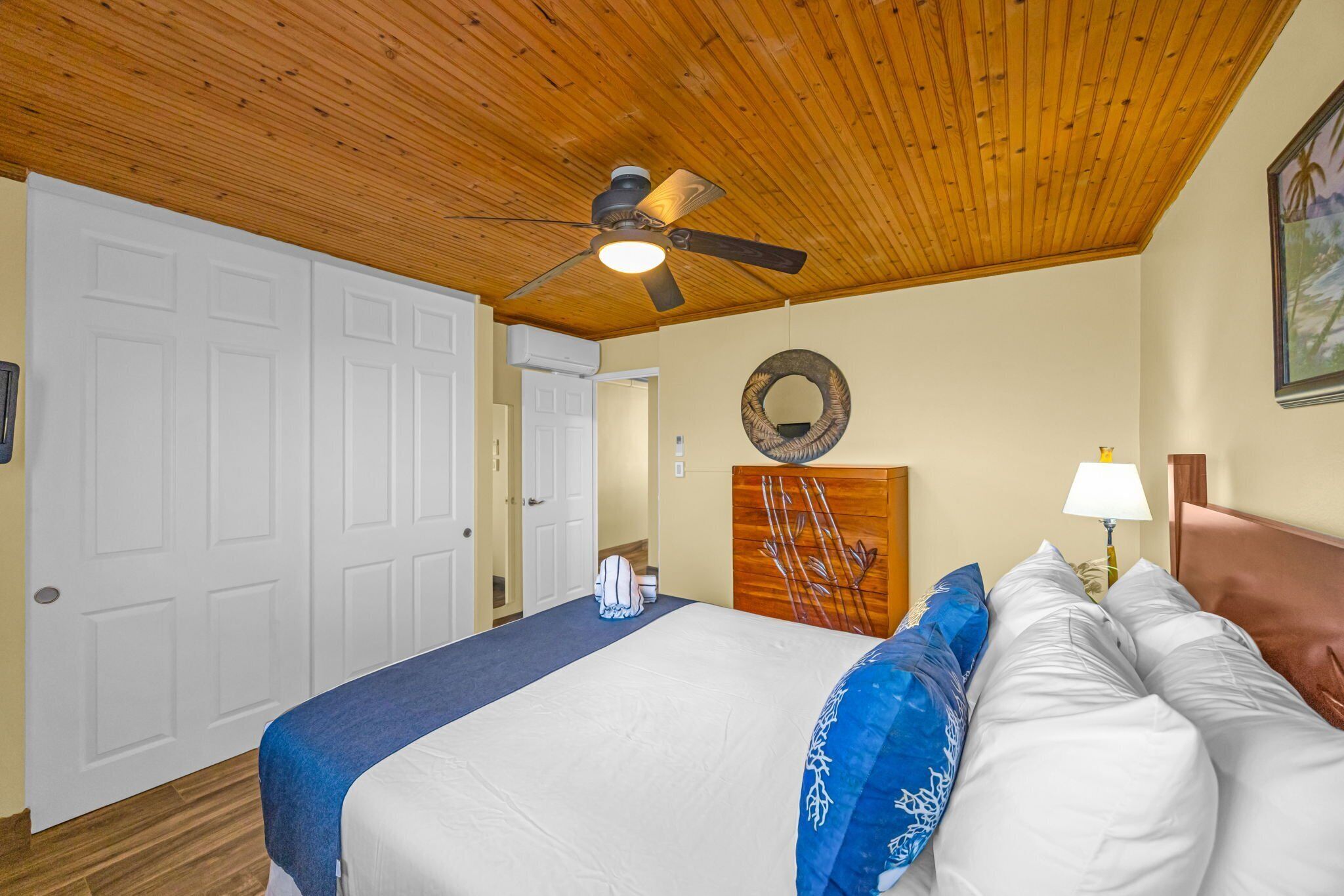 Un dormitorio con techo de madera, cama con ropa de cama blanca y azul, ventilador de techo y un espejo fijado a la pared.