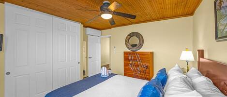 Un dormitorio con techo de madera, cama con ropa de cama blanca y azul, ventilador de techo y un espejo fijado a la pared.