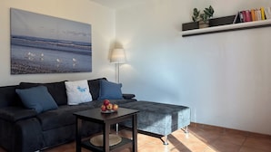 Una sala de estar con un sofá oscuro, una mesita con manzanas, una lámpara y una estantería con libros y plantas.
