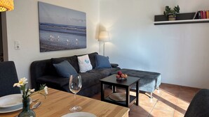Un salón con sofá, una mesa de centro, un cuadro de pájaros en la pared, una lámpara y una estantería con libros y una planta.