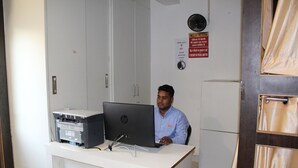 Un homme est assis à un bureau, devant un ordinateur et une imprimante, avec un meuble en arrière-plan.
