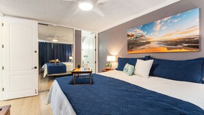 Une chambre avec un grand lit, un ventilateur de plafond, un tableau représentant un coucher de soleil et une vue sur la salle de bain.