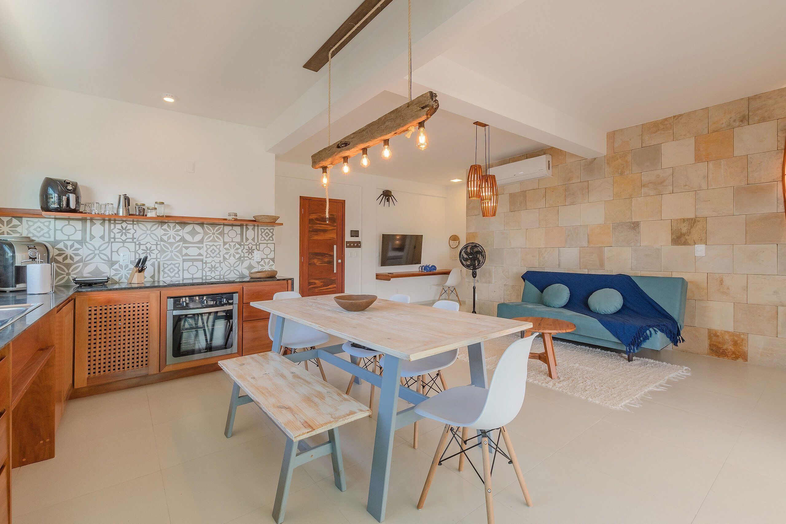 A modern kitchen with wooden cabinets, a dining table with chairs, and a blue sofa in a living area with a stone wall.